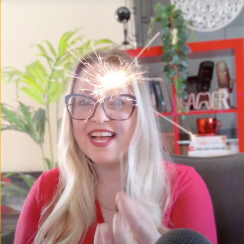 Gina Dunn, brand strategist and Morning Cup of Brand Spark host, smiling and holding a sparkler, wearing glasses and a red