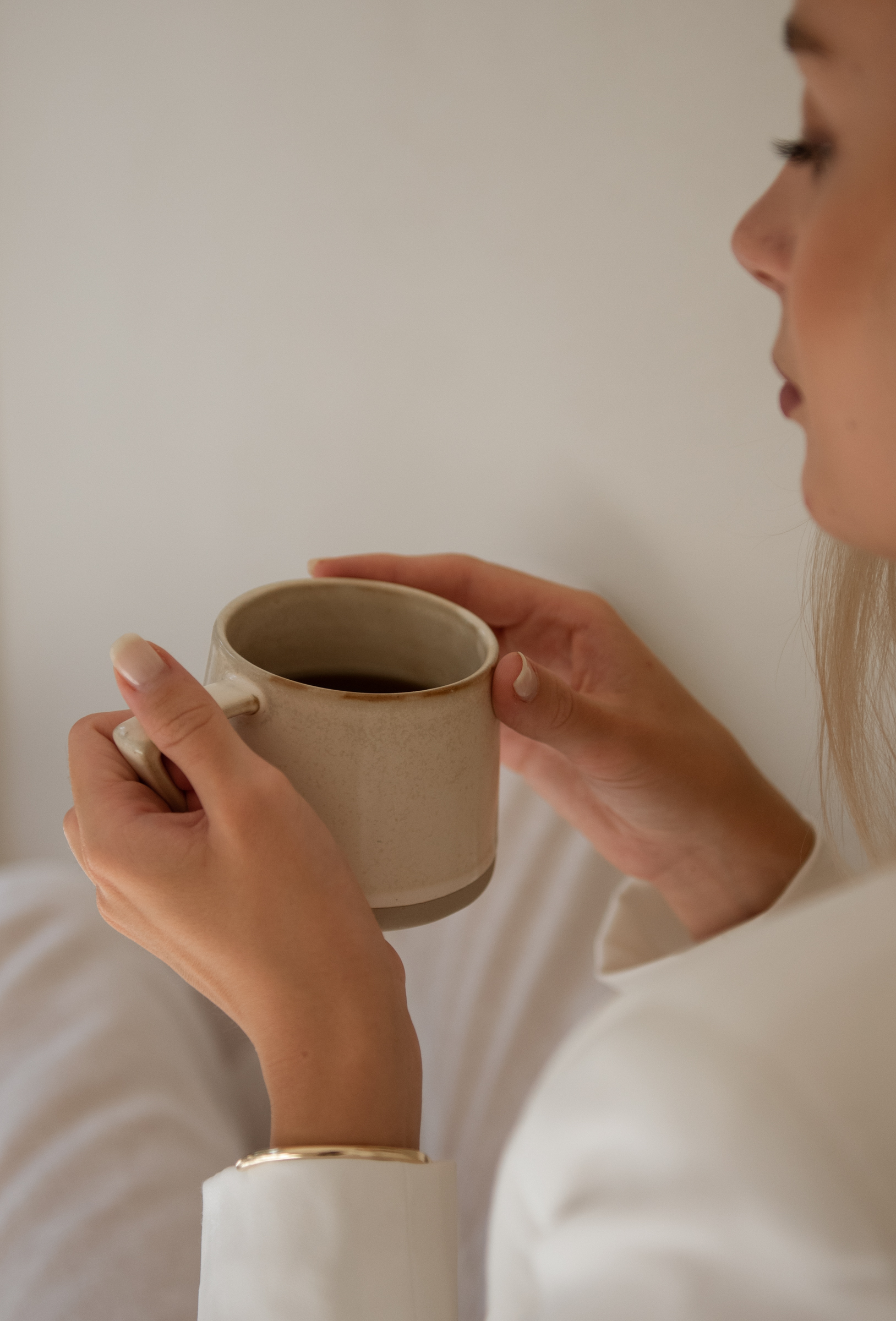 Woman in white top holding a rustic ceramic mug with both hands, gold bracelet visible, evoking a calm morning brand clarity