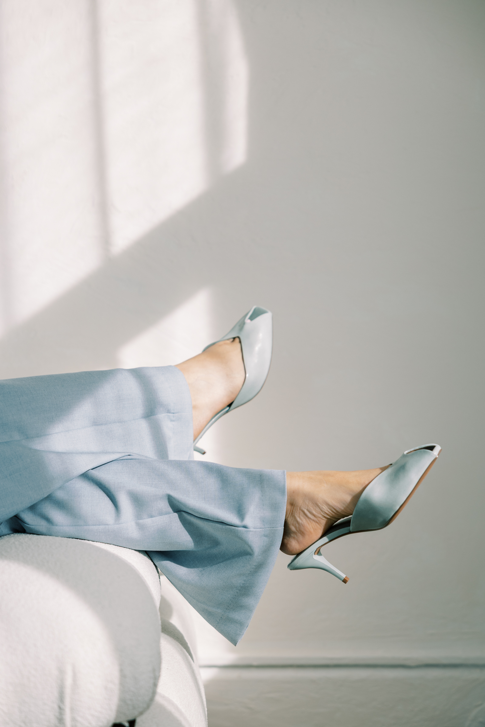 Legs crossed on a white chair wearing light blue wide-leg trousers and pale blue kitten heel mules against a softly lit